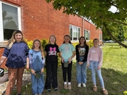 Six 5th grade girls stand in a line against a red brick building under the shade of a tree. Several of them are holding small green plastic pots, ready for a gardening or planting activity.